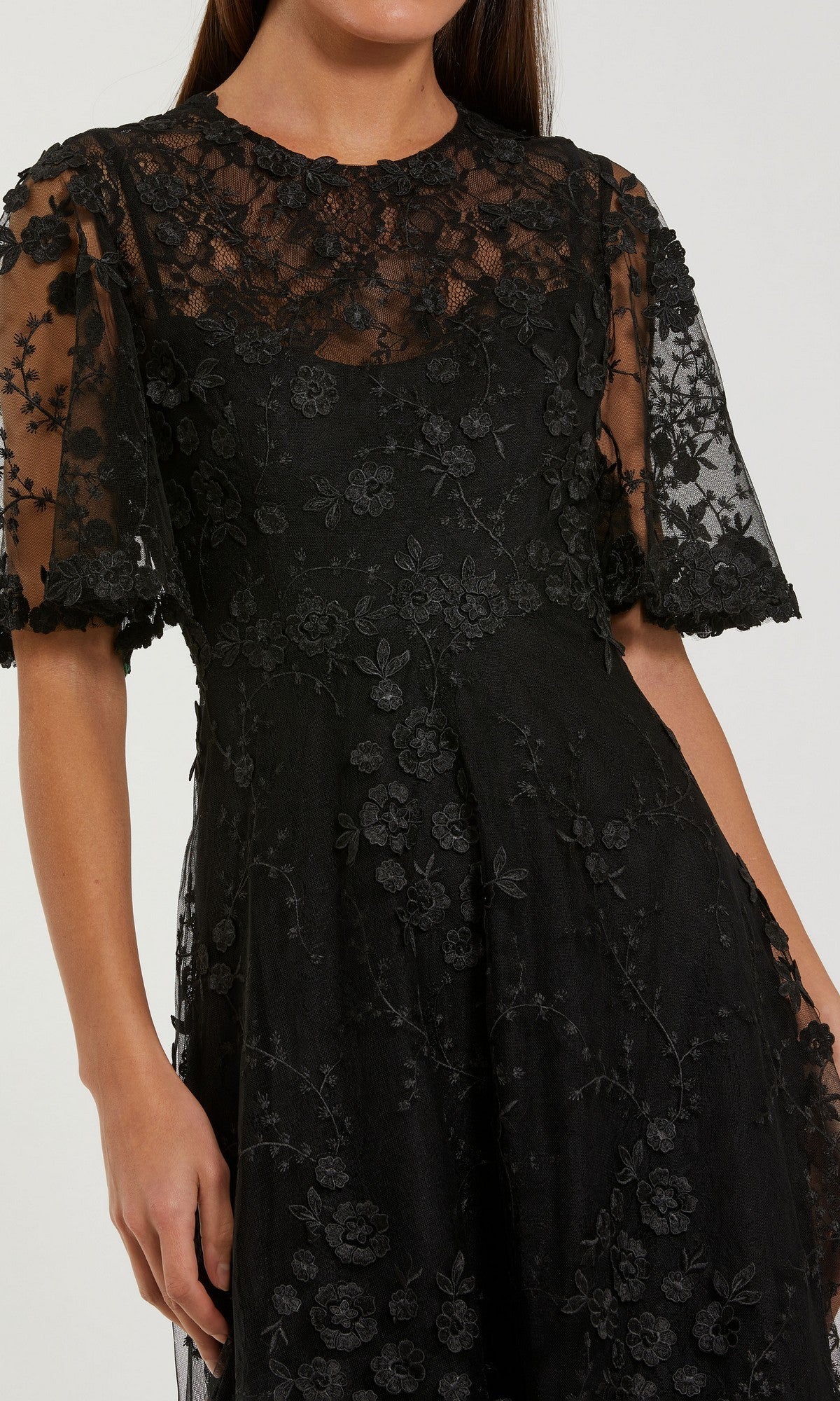 Woman in a black Mac Duggal 69036 Midi Wedding Guest Dress featuring floral embroidery and sheer short sleeves, shown from shoulders to mid-thigh against a plain background.